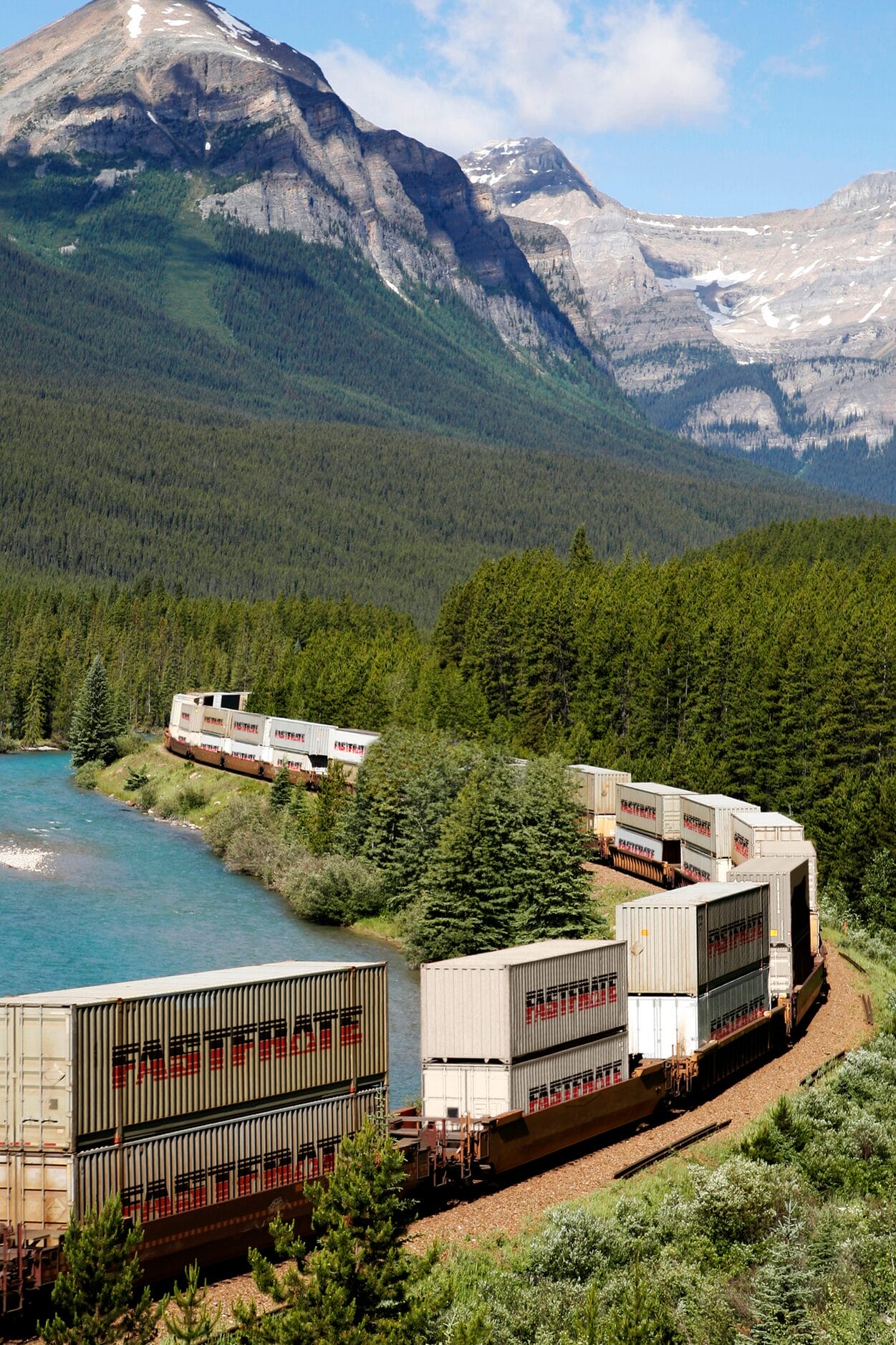 Train with cargo containers curves along a river in a mountainous landscape