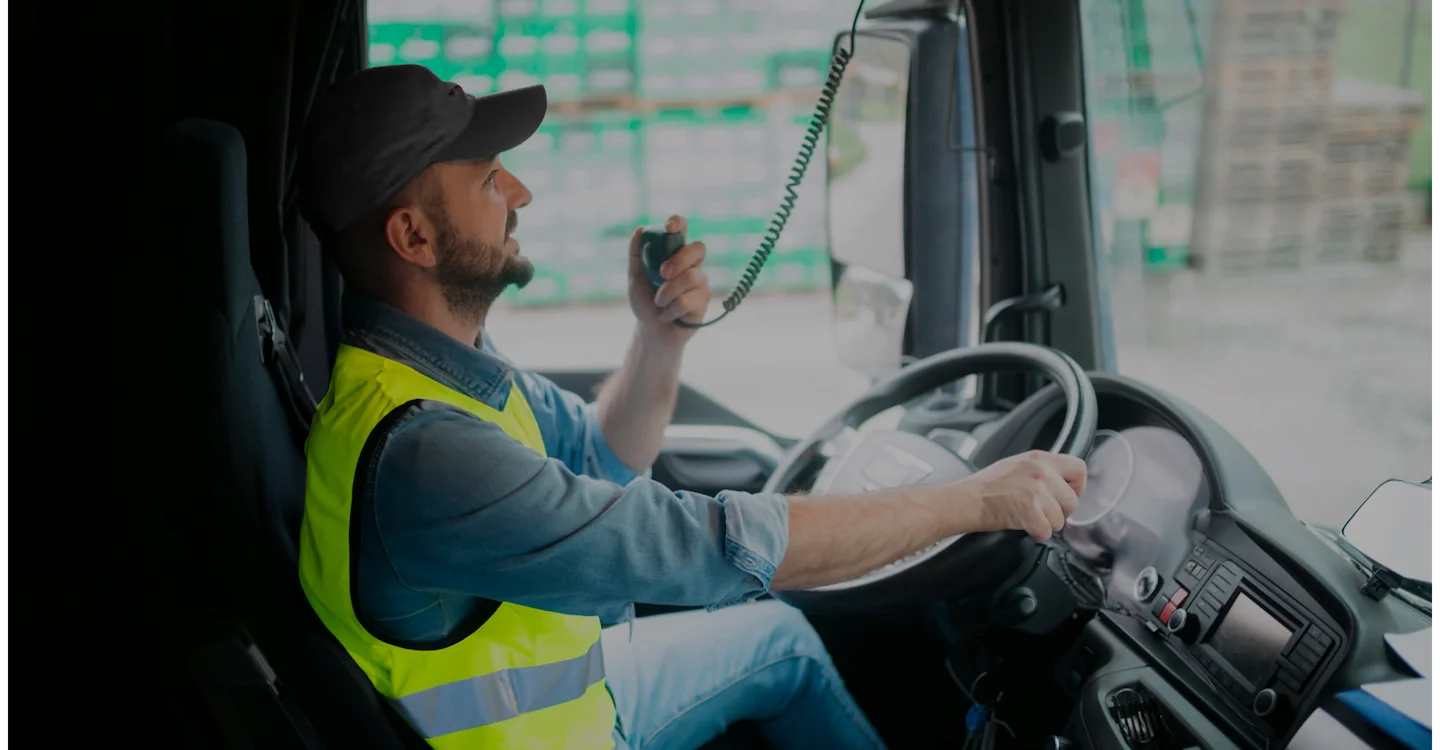 Truck driver wearing a safety vest, speaking into a radio inside the cab