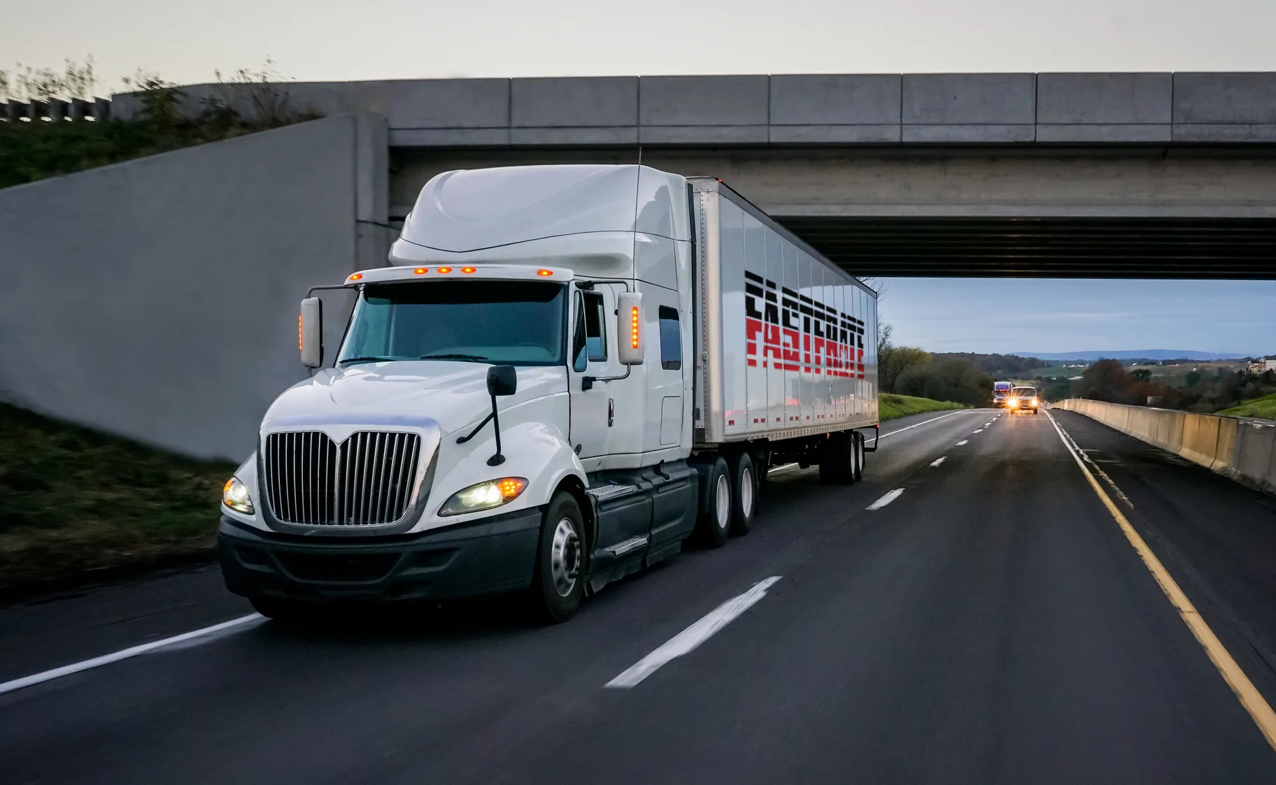 Large white truck driving on a highway under an overpass at dusk