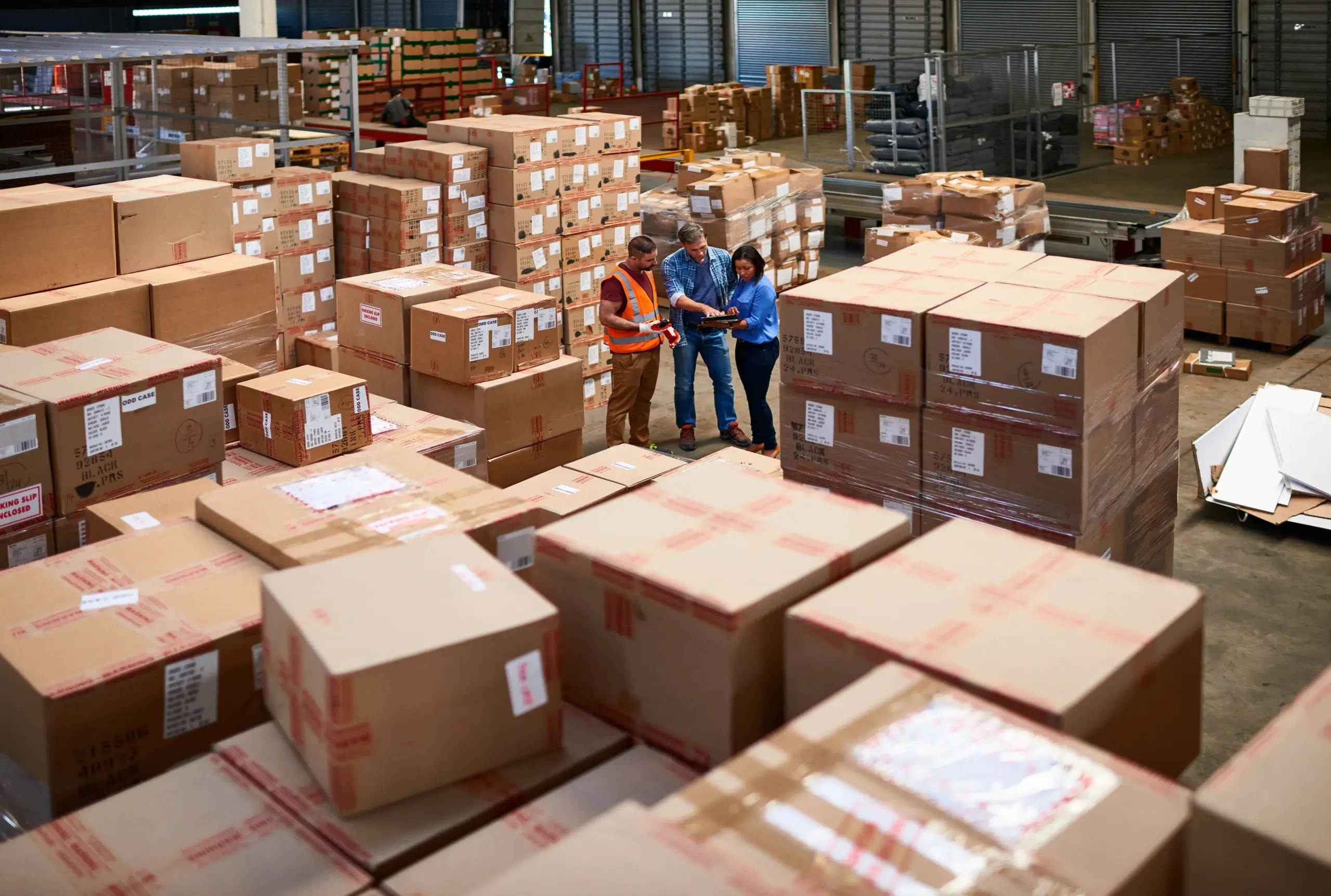 Workers discussing logistics among stacked cardboard boxes in a warehouse
