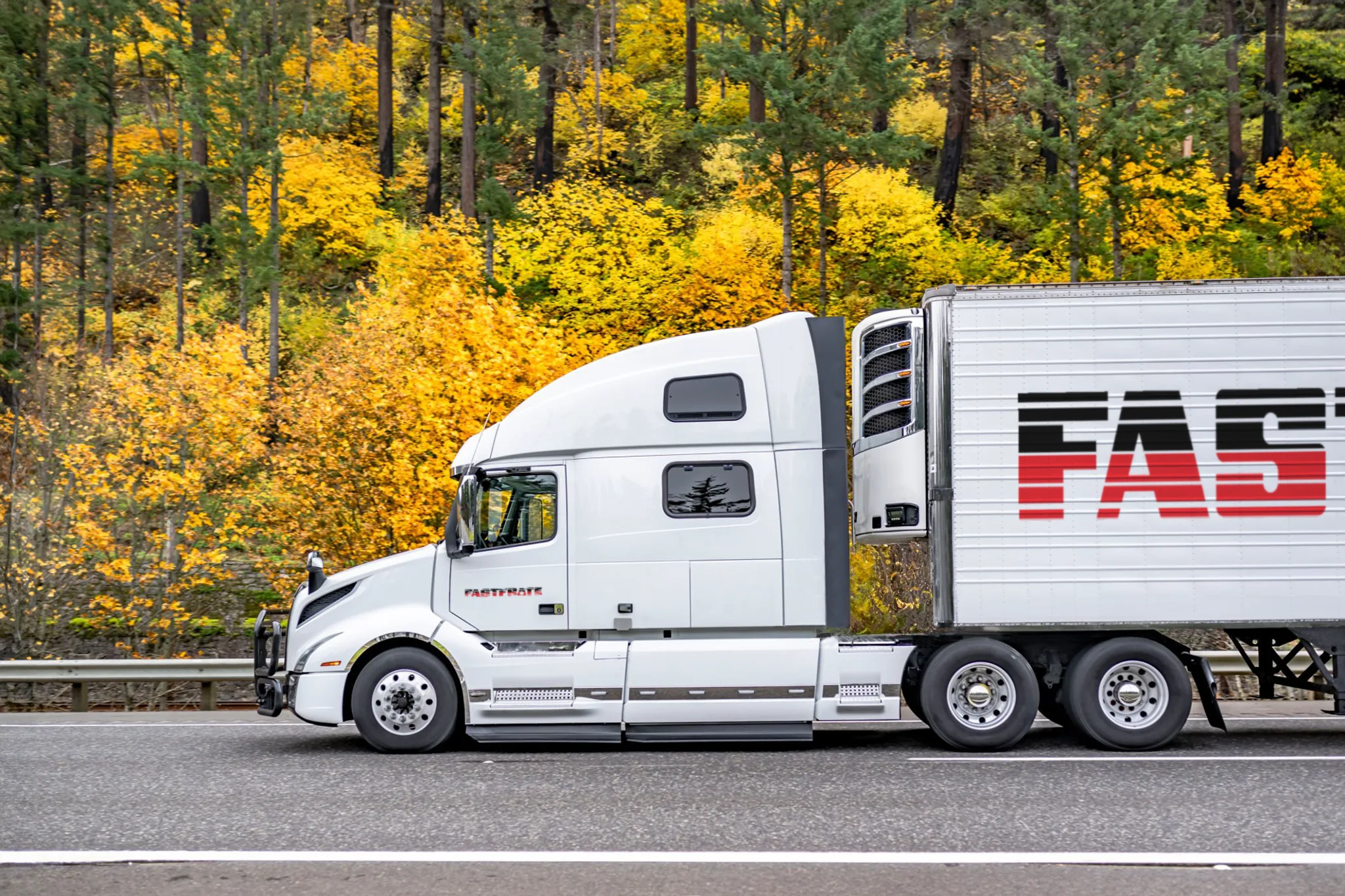 White semi-truck with a "FAST" trailer on a highway surrounded by autumn foliage