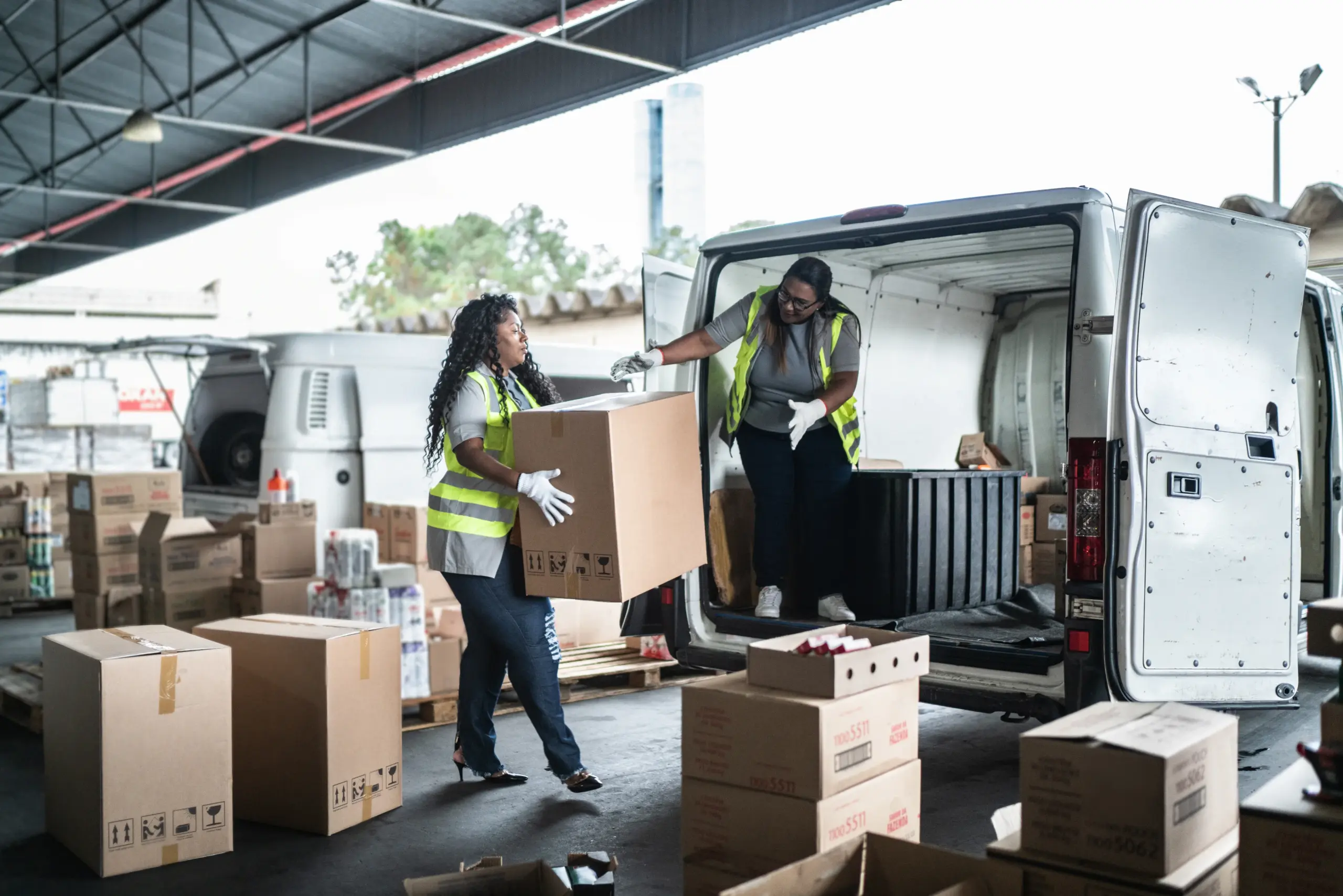 Two workers in vests loading boxes into a delivery van at a warehouse