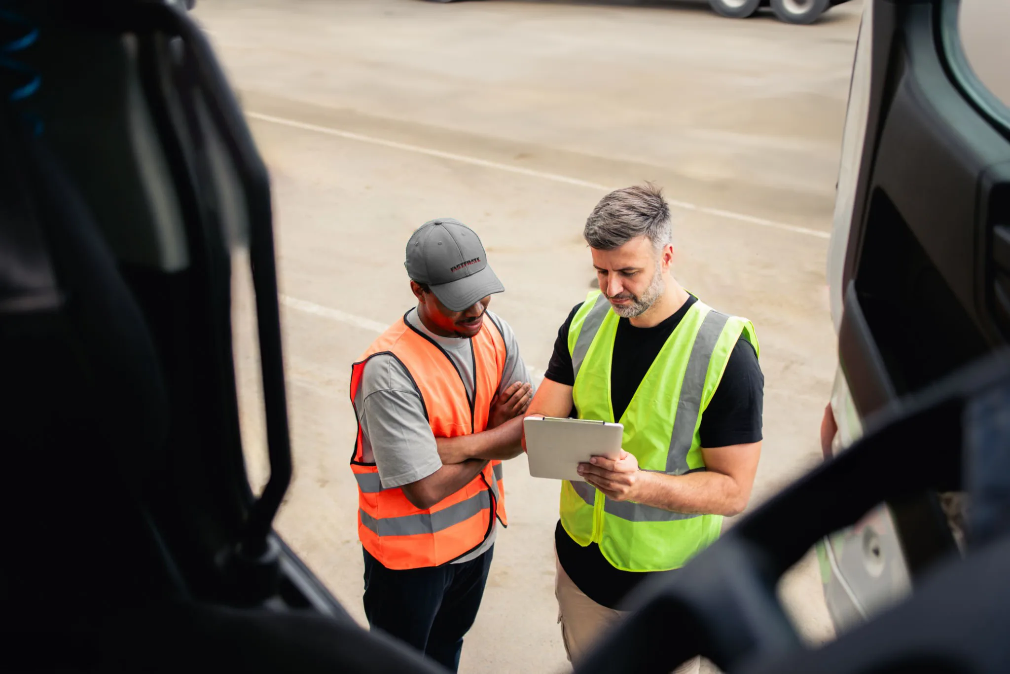 Two workers in safety vests discussing a tablet outdoors near trucks