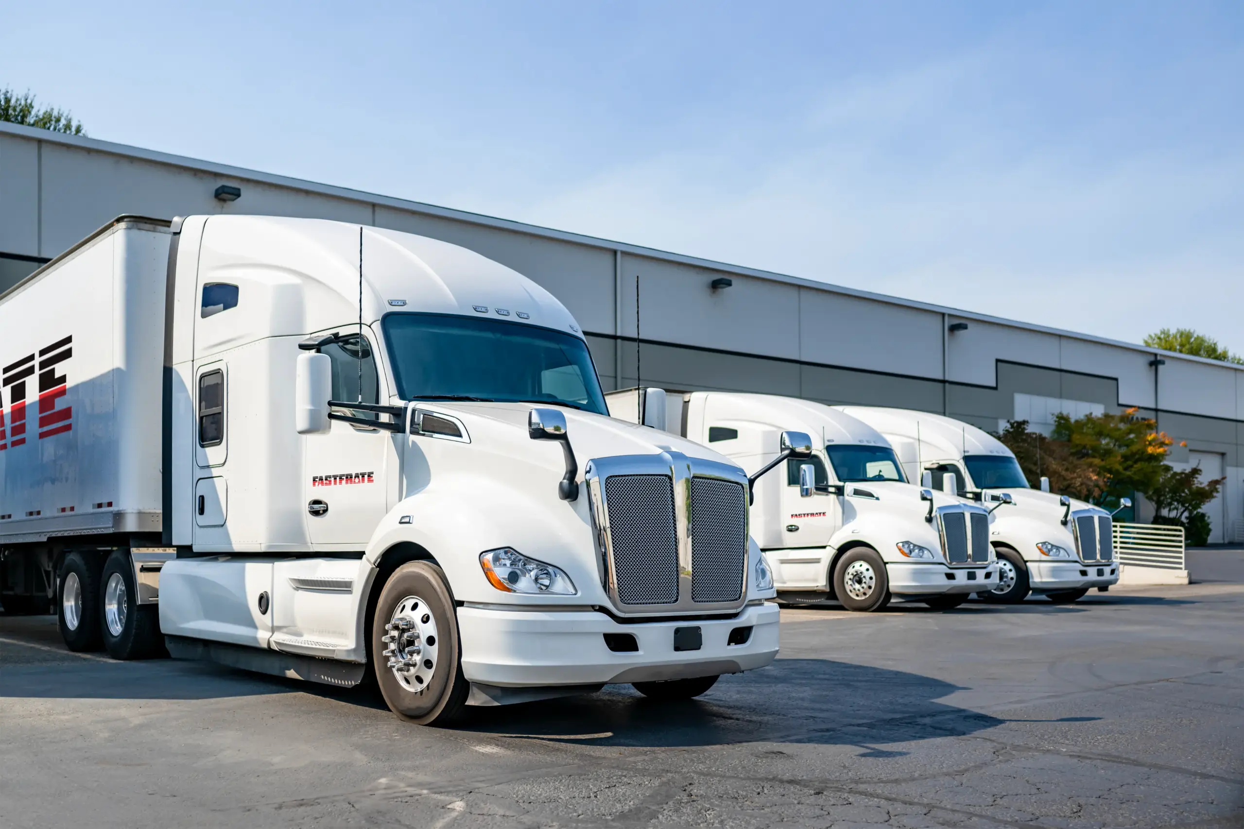 Three white semi-trucks parked outside a warehouse on a sunny day
