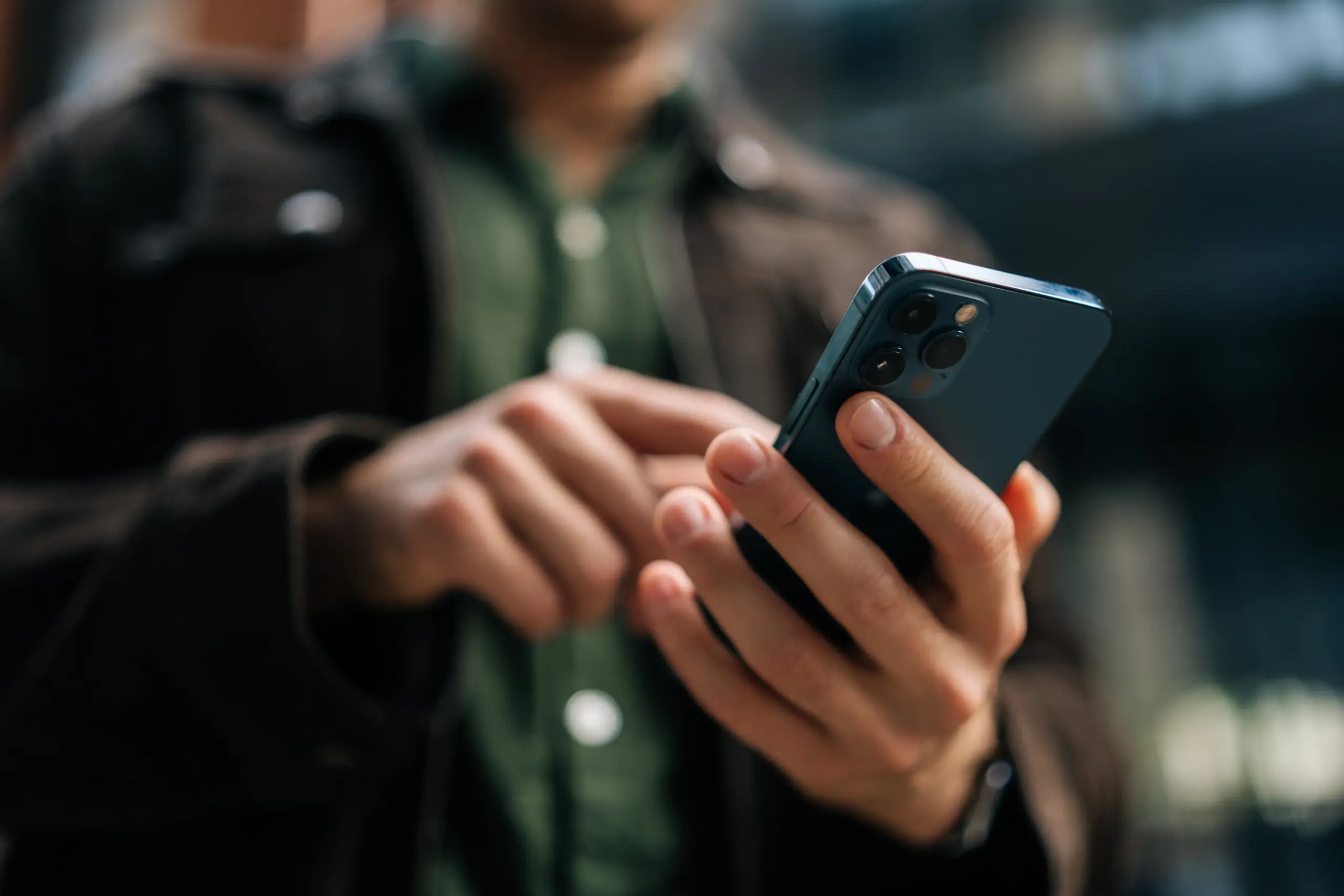 Person using a smartphone while standing indoors, wearing a dark jacket and green shirt